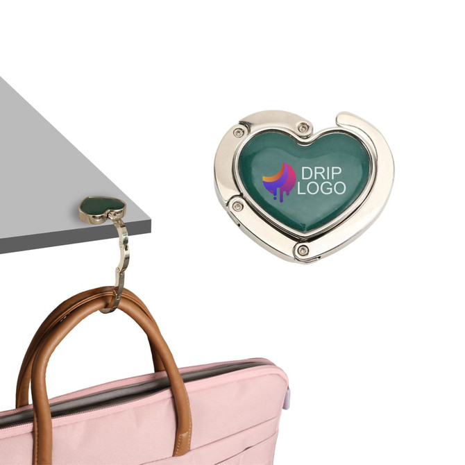 A heart-shaped foldable desk hook in silver and green, supporting a handbag on a table edge, featuring a logo.
