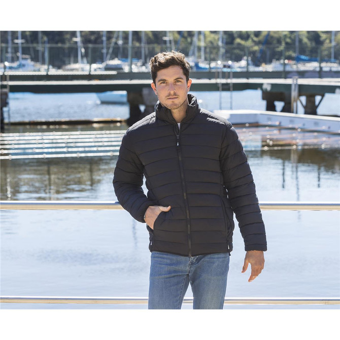A unisex black puffer jacket with a zip front and side pockets, modelled by a man beside a waterfront.