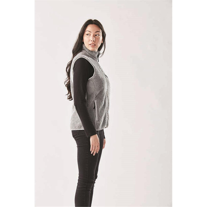 A side view of a women's grey sherpa fleece vest, worn over a black long-sleeve top, with a logo visible.