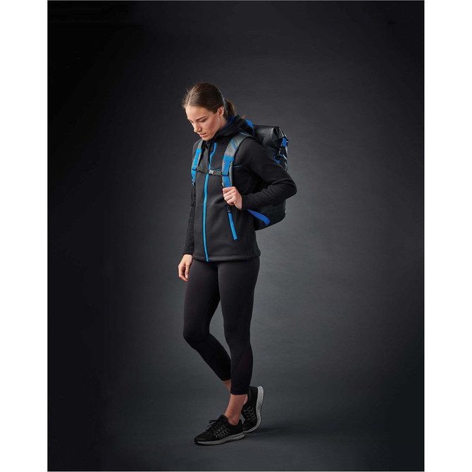 A woman in a black hoodie with blue accents, wearing black leggings and a backpack, stands against a dark background.