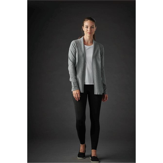 A woman wearing a grey Soho cardigan over a white top and black leggings, standing against a dark background. The cardigan has a logo.