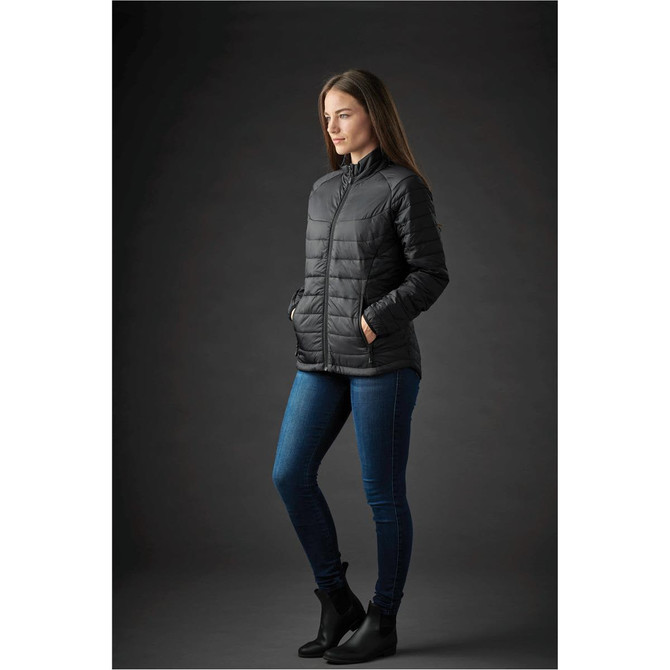 A woman wearing a black puffer jacket with a zip, paired with blue jeans and ankle boots. The jacket features a logo.