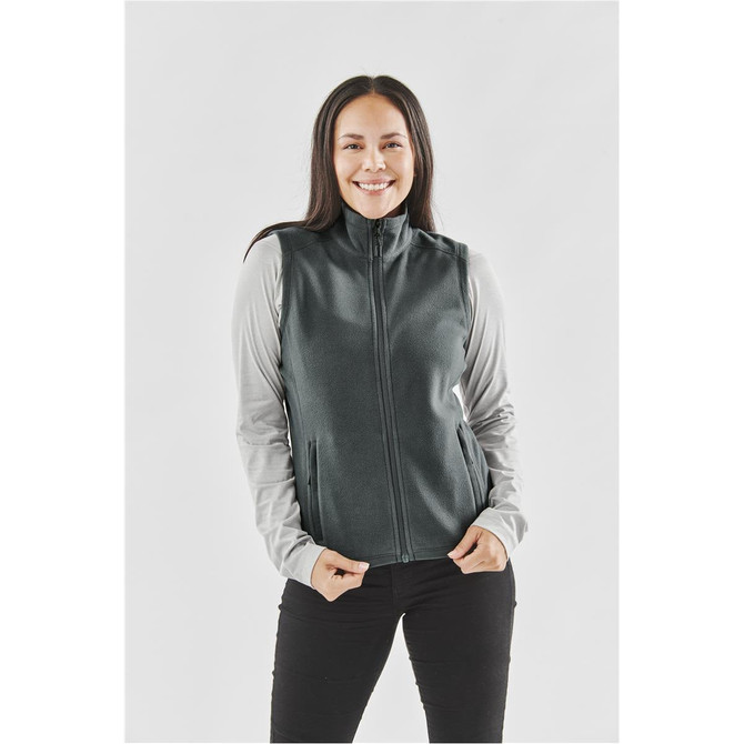 A woman stands wearing a dark green fleece vest over a light grey long-sleeve top, smiling at the camera.