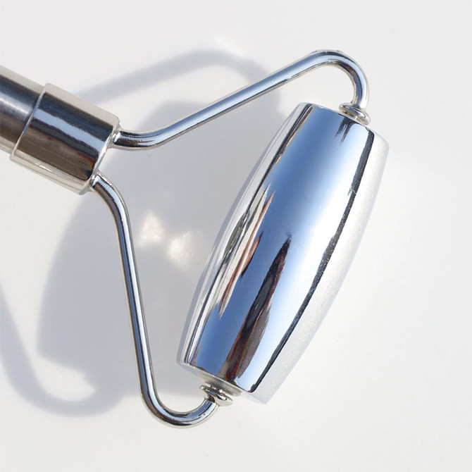 A stainless steel facial roller with a shiny, reflective surface and a curved design. It has a logo on the handle.