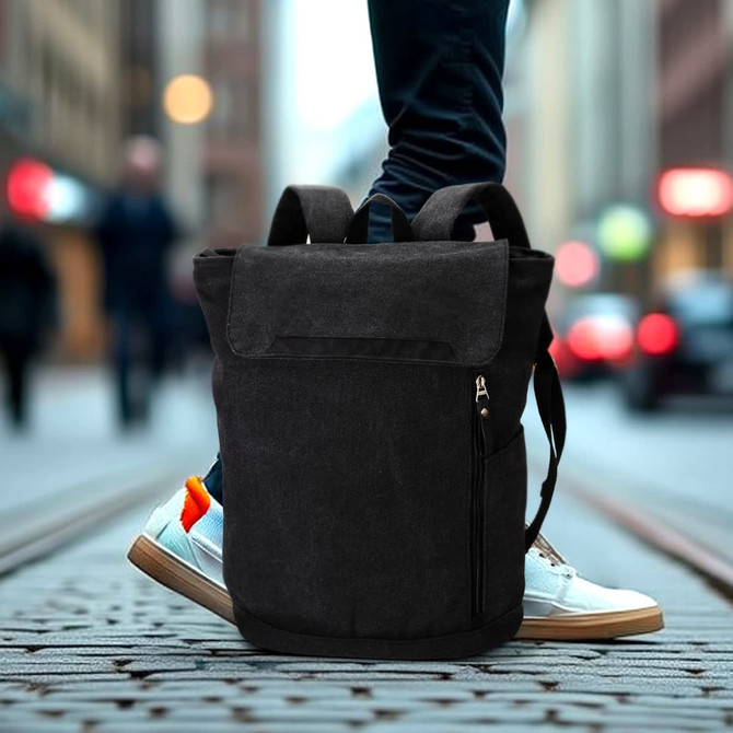 A black canvas backpack with a zippered pocket, placed on a cobblestone street, with a person's feet in white sneakers nearby.