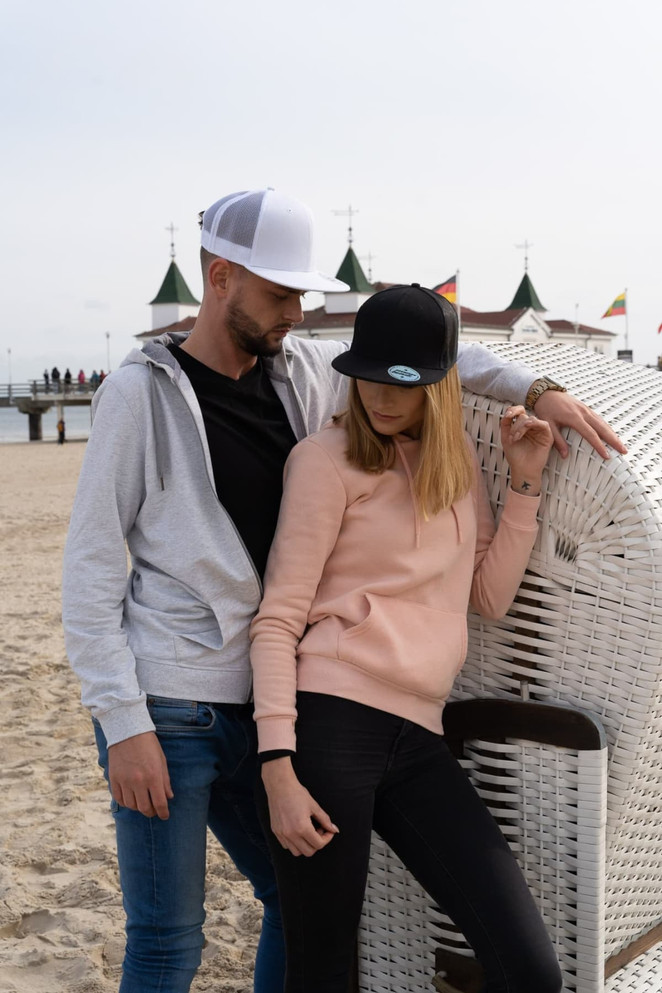 A couple wearing casual clothing, featuring a grey hoodie and a pink sweatshirt, along with black and white snapback caps.