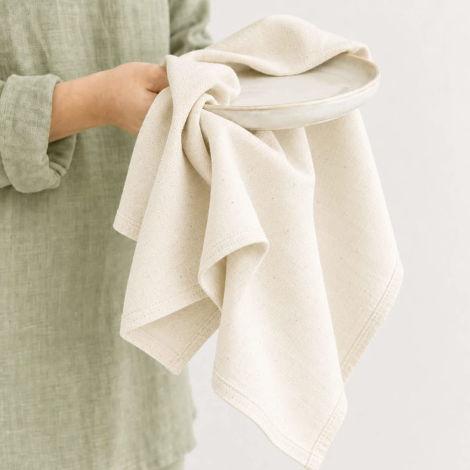 A medium cotton tea towel in a natural beige shade held over a ceramic plate, featuring a subtle texture.