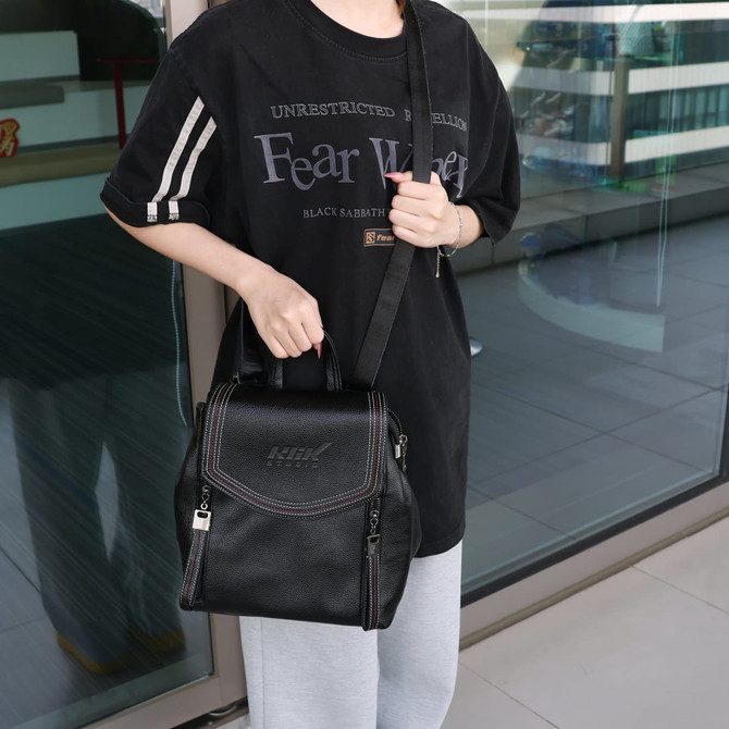 A black convertible leather backpack is held by a person wearing a black T-shirt and grey pants. The backpack has a logo.