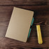 An A5 notebook in a brown cover with a blue and green pen beside a yellow highlighter on a wooden surface.