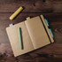 A brown A5 notebook with lined pages, accompanied by a green pen, a blue pen, and a yellow highlighter on a wooden surface.