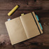 An A5 notebook with lined pages, featuring a yellow highlighter, sticky notes, and pens placed on a wooden surface.