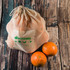 A reusable mesh produce bag in natural color, with two oranges placed beside it. The bag features a logo.