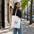 A woman carries a cream-coloured long handle cotton tote bag with a logo, walking along a city street.