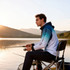 A man wearing a blue and white polyester fishing hooded jacket sits by a lake, fishing at sunset.
