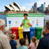 A man in a green and white polo shirt stands before a recycling information display, surrounded by a group of people and bins.