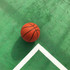 An orange basketball resting on a green court with white lines. The ball has a textured surface and seams.