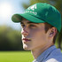 A green cap with a logo is worn by a young man, set against a blurred outdoor background.