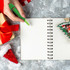 A green tree-shaped stress reliever pen held above a blank spiral notebook, surrounded by a gift and festive decorations.