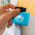 A turquoise magnetic clip is held against a surface, featuring a logo on the front and a black handle.