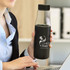 A drink bottle with a black silicone sleeve and transparent glass upper section, featuring a logo.
