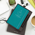 A teal Moleskine notebook and pen gift set in a brown box, with office supplies and a plant in the background.