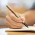 A hand holding a Kraft Paper Pen, with a black nib, writing in a notebook. The pen has a logo printed on it.