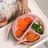 A pink kids' plate set with compartments holds sliced carrots, broccoli, and sauce-covered pasta; accompanied by a fork and spoon.