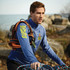 A man wearing a blue quarter zip jumper, with a logo and harness, is outdoors near water and rocks.