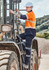 A man wearing an orange and navy shirt and dark work pants climbs into a large machinery vehicle on a worksite.
