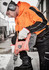 A worker in an orange and black hi-vis pullover hoodie using a power drill while wearing protective gear and gloves.