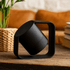 A black wireless speaker with a unique geometric design, set against a decorative background with plants and books.