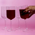 Four clear glass wine glasses with ribbed design, displayed against a pink background, one held with red liquid.