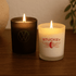 Two candles on a wooden surface: one black and one white, both with lit flames. The white candle has a logo.
