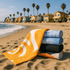 A stack of five bath towels in various colours, including yellow, blue, and black, on a sandy beach with palm trees and houses in the background.