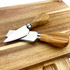 A wooden cheeseboard featuring two stainless steel cheese knives with wooden handles.