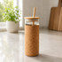 A drink bottle with a glass body, cork exterior, bamboo lid, and a straw in a bright kitchen setting.