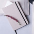 A beige A5 cotton notebook with a black elastic strap and a pink flower embellishment on the cover.
