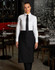 A woman in a black short waist apron over a white shirt and tie, standing in a restaurant setting.