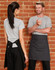 A grey canvas short waist apron in black worn by a person facing another person in casual attire against a brick wall.