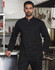 A black men's functional chef jacket, featuring a sleek design and a prominent logo, displayed in a kitchen setting.