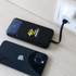 A black power bank with a logo is charging near a phone, connected to a wall socket with a cable.