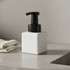A white square foaming soap dispenser with a black pump, placed on a grey surface next to a textured cloth.