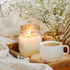 A white scented candle with a warm glow is positioned beside a cup of coffee and a vase of flowers on a textured tray.