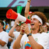 A man with curly hair joyfully holds a red and white megaphone, surrounded by cheering fans in a sports setting.