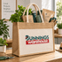 A jute beach bag in natural colour with cotton handles and a front pocket, featuring a logo, filled with gardening items.