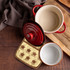 A large quilted pot holder in a cream and tan design, featuring a red ceramic pot and a white bowl on a wooden surface.