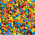 Colourful scattered M&Ms in various shades, including blue, red, green, yellow, and brown, with a brand logo visible.