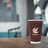 A brown compostable paper cup with a double wall design, featuring a leaf logo, placed on a wooden table.
