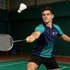A young man wearing a navy and teal polo shirt with a logo plays badminton, swinging a racket at a shuttlecock.
