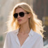 A pair of black classic aviator sunglasses worn by a model with blonde hair, set against a blurred outdoor background.